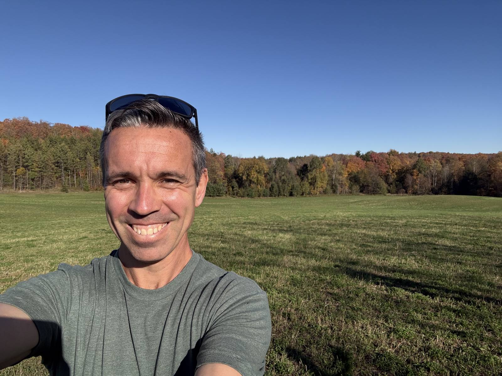Trevor smiling in an open field with autumn trees and a clear blue sky behind